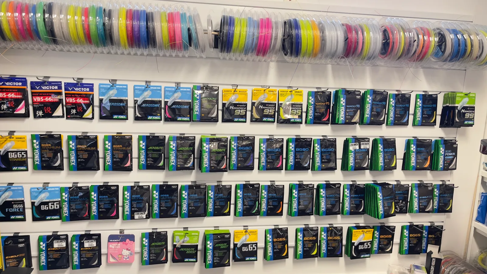 A wall of Yonex badminton string — reels of coloured string hanging on a rail above rows of packeted string including BG80, Nanogy 95, Aerobite and BG65.